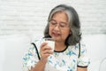 Asian Senior woman holding glass milk while relaxing on a sofa living room for retirement wellness. Elderly Woman drinking a glass