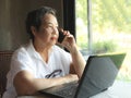 Asian senior woman sitting with computer laptop on table , talking on the phone, looking away out of the window Royalty Free Stock Photo