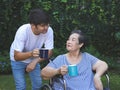 Asian senior woman sitting on wheelchair, drinking coffee or tea with her son in the garden. smiling happily Royalty Free Stock Photo