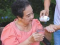 Asian senior woman sitting on wheelchair holding glass of water, get medical pill from her son. garden, outdoor Royalty Free Stock Photo