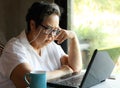 Asian senior woman sitting by the windor with blue cup of coffee and computer laptop on table , looking thoughfully at computer Royalty Free Stock Photo