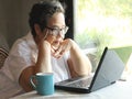 Asian senior woman sitting by the window with blue cup of coffee and computer laptop on table looking at computer laptop ,chin on Royalty Free Stock Photo