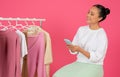 Asian shopper lady with smartphone sitting contemplatively near clothing rack,