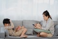 An Asian single mother is sitting on the sofa in the living room of the house using a tablet with her son happily playing games