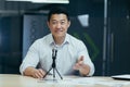 Asian smiling young man sitting in office at desk in front of camera, talking on microphone via video call, blogging Royalty Free Stock Photo