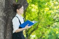 Asian student reading blue book, against green of summer park.