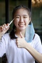 Asian teenager toothy smile with protection mask hanging beside head and themometer in hand Royalty Free Stock Photo