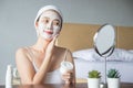 Asian woman applying moisturizer face scrub peeling white clay mask on skin, looking in mirror with smiley face using cream