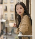 Asian woman in balcony thinking - outdoors lifestyle portrait of young beautiful and thoughtful Japanese girl at drinking morning