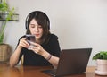 Asian woman in black shirt wearing headphones, sitting at wooden table with computer laptop, using mobile phone. white wall with Royalty Free Stock Photo