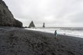 Asian woman with blue jacket, shorts and backpack standing on black beach, looking towards the extreme waves of Reynisfjara