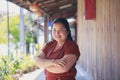 An Asian woman in a brown shirt crossing her arms looking at a camera next to wooden wall