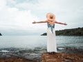 Asian woman in dress and straw hat looking away to the turquoise calm sea water. female open arms on the rock at tropical beach in Royalty Free Stock Photo