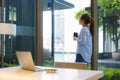 Asian woman drinking coffee in mug and smiling happily while taking a break from work at her computer during work at home in the Royalty Free Stock Photo