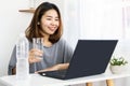 Asian woman drinking fresh water at work place sitting at office hand typing on laptop and holding glass of water