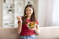 Asian woman eating vegetable salad while sitting on couch in living room Royalty Free Stock Photo