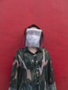 An asian woman with a face mask and face shield looks at a camera. Agaisnt a red wall.