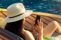 Asian woman with hat and swimsuit sitting in chair at poolside and using smartphone on summer vacation. Luxury life. Checking News