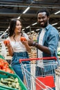 Asian woman holding fresh tomatoes near african american man showing thumb up in supermarket