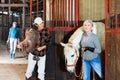 Asian woman holding saddle while elderly female stable keeper leading horse out of stall