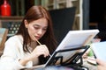 Asian woman holds a paintbrush in right hand as dominant hand, left hand is on laptop previewing illustration while drawing along Royalty Free Stock Photo