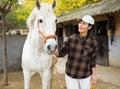 Asian woman horse breeder leading white racehorse along stables outdoors Royalty Free Stock Photo