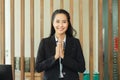 Asian woman hotel receptionist smiling and hand up wai thai style greetings hand to pay respect standing at front of lobby.