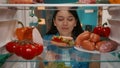 An Asian woman looks into the refrigerator, examines its contents. A woman, pulling up a plate with a sandwich, inhales