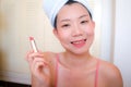 Asian woman and makeup - young happy and beautiful Chinese girl with towel head wrap applying lipstick looking at bathroom mirror Royalty Free Stock Photo
