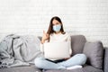 Asian woman in mask sits on sofa with laptop in living room