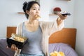 Asian woman with pizza in mouth eating unhealthy junk food sitting in bed with potato chips another hand holding TV remote Royalty Free Stock Photo