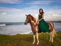 Asian woman riding horse near the ocean. Outdoor activities. Woman wearing long green dress. Traveling concept. Cloudy sky. Copy Royalty Free Stock Photo