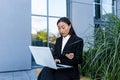 Asian woman shopping sitting on a bench near an office building, using a laptop and credit card, online sales