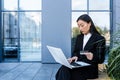 Asian woman shopping sitting on a bench near an office building, using a laptop and credit card, online sales, making choices