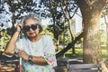 Asian woman sits in a wooden chair with a cane, is blind and wearing black glasses as she sits alone in a park.