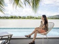 Asian woman sitting on beach chair near swimming pool under palm tree. infinity edge pool with river view. Royalty Free Stock Photo