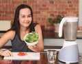 Asian woman sitting in kitchen  preparing healthy smoothie , holding bowl of gree vegetable in her hand smiling to camera Royalty Free Stock Photo