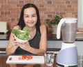 Asian woman sitting in kitchen  preparing healthy smoothie , holding bowl of green vegetable in her hand smiling to camera Royalty Free Stock Photo