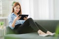 Asian woman sitting on sofa at home using digital tablet for work or online shopping. Touch screen freelance device for business o Royalty Free Stock Photo