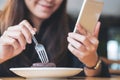 An Asian woman with smiley face holding and using smart phone while eating brownie on wooden table Royalty Free Stock Photo