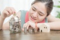 Asian woman smiling and holding coin above her jar glass bank next to house model and stacked of coins with tree house decoration