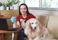 Woman smiling and looking at camera , holding blue cup of coffee,  sitting on couch in living room with computer in front of her Royalty Free Stock Photo