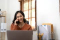 Asian woman talking on her mobile phone and using laptop while sitting at desk and working from home. Home office. Royalty Free Stock Photo