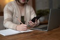 An Asian woman using her smartphone and taking notes on paper while sitting at a table in a cafe Royalty Free Stock Photo