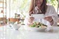 An asian woman using smartphone to taking photo of salad in a white plate on table in cafe