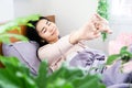 Asian woman waking up in bed in morning stretching her arms with smiling face happy with fresh green plants in bedroom