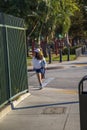 An Asian woman walking along a sidewalk surrounded by a green metal fence and lush green trees and grass in Pasadena California