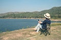 Asian woman wear hat and sitting relax on portable chair and playing smartphone nearly lake at National Park.