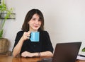 Asian woman wearing black shirt, sitting at wooden table with computer laptop and plant pot, holding blue coffee cup, smiling and Royalty Free Stock Photo