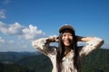 An Asian woman wearing a casual dress and a white cap smiling happily with a mountain background, forests, clouds and skies